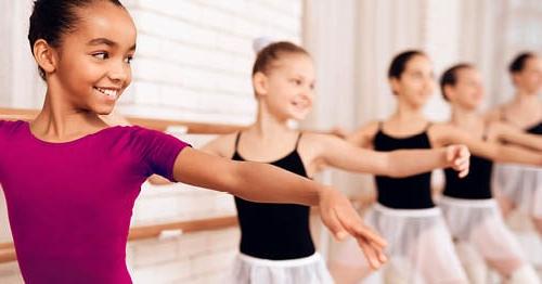 A group of young female ballet students practicing at the barre. One is wearing pink and the rest are wearing a black leotard.